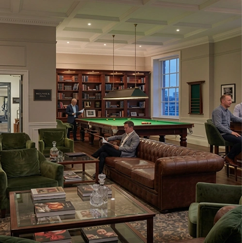 Het luxe interieur van een moderne lounge, met een comfortabele leren bank en groene fauteuils rond een glazen salontafel. In de achtergrond is een brandende open haard en een pooltafel te zien.